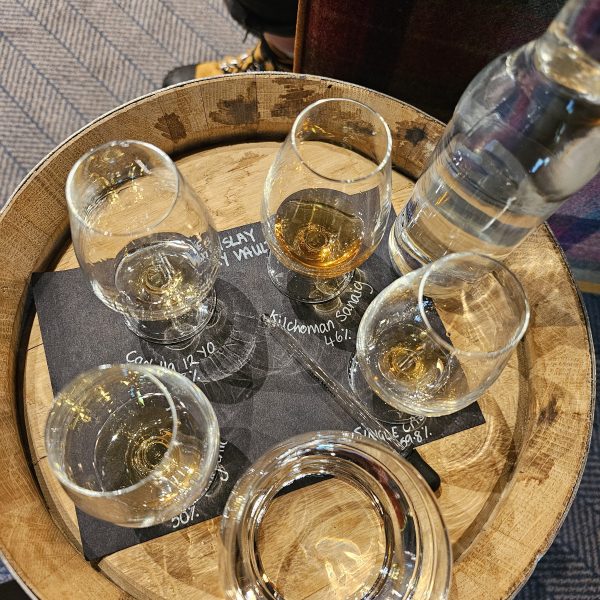 Five whiskey glasses on a wooden barrel, with a water bottle and a person in background.