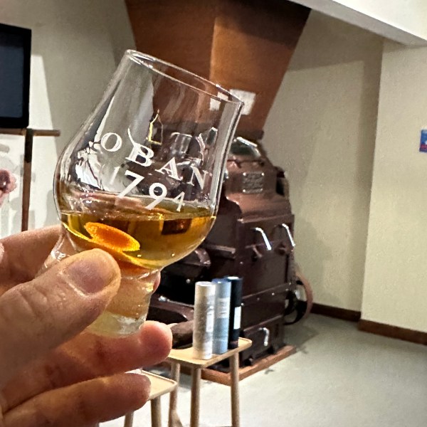 Hand holding a glass of Oban 1794 whiskey with distillery equipment in the background.