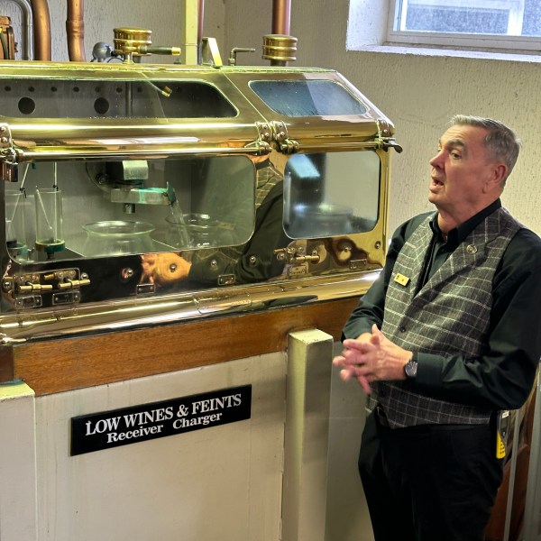 Man explaining a distillery machine labeled 'Low Wines & Feints' in an industrial setting.