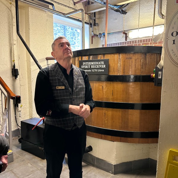 Man in vest stands by large wooden barrel labeled 'Intermediate Spirit Receiver' in an industrial room.
