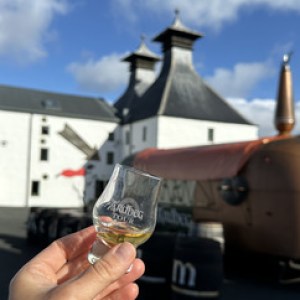 Hand holding a whiskey glass with distillery building in the background.
