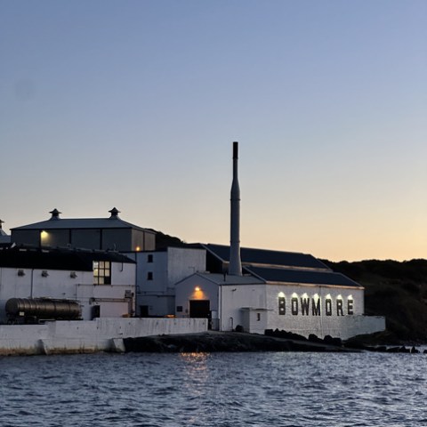 White distillery building beside the sea at dusk with 'BOWMORE' sign illuminated.