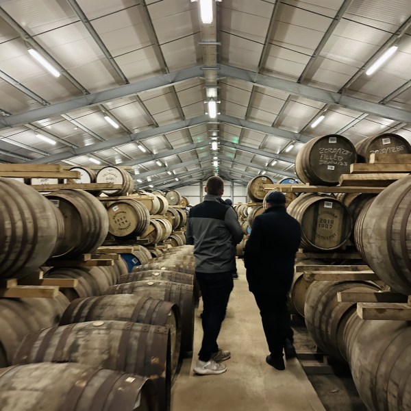 Two people walking in a warehouse filled with stacked wooden barrels.