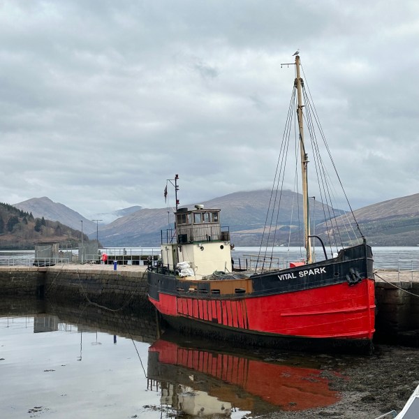 Red and black boat named 'Vital Spark' docked beside stone wall, with mountains in the background.