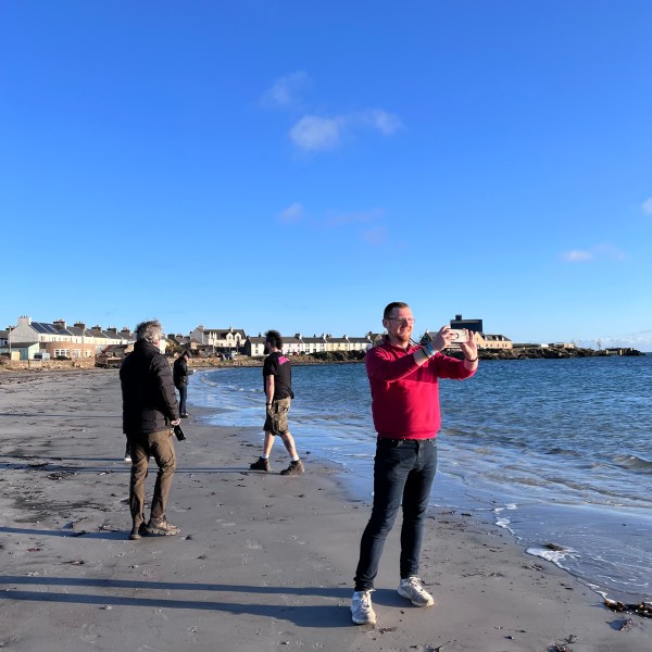 People on a sandy beach, one in red taking a photo, with houses and ocean in the background.