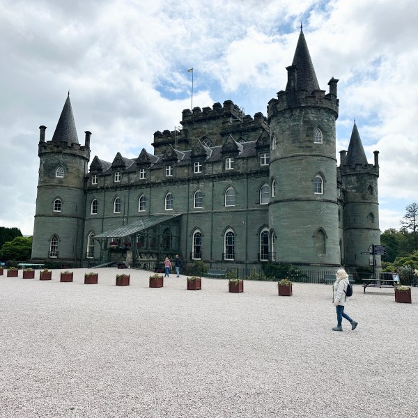 Stone castle with round towers, cloudy sky; person walking in foreground.