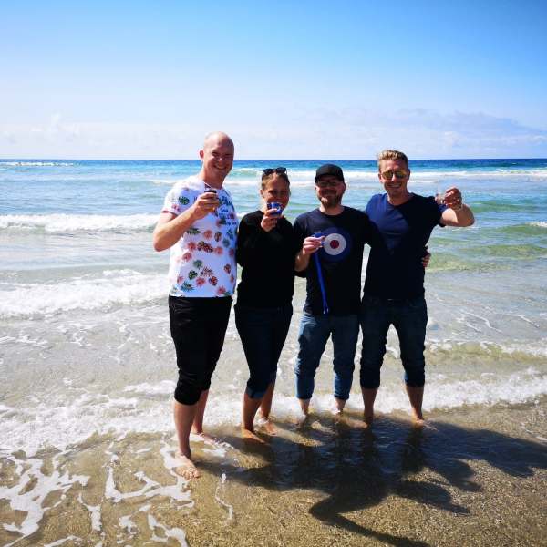 Preview of our four day islay tour. Four friends standing on a beach with drinks.