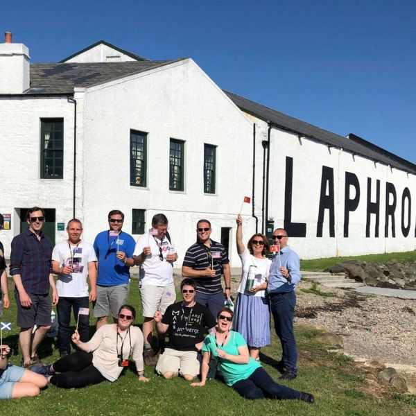Group of people posing in front of a Laphroaig distillery building under a clear blue sky.