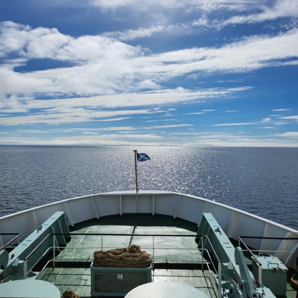 Ship's bow with ropes and flag on open sea under cloudy sky.