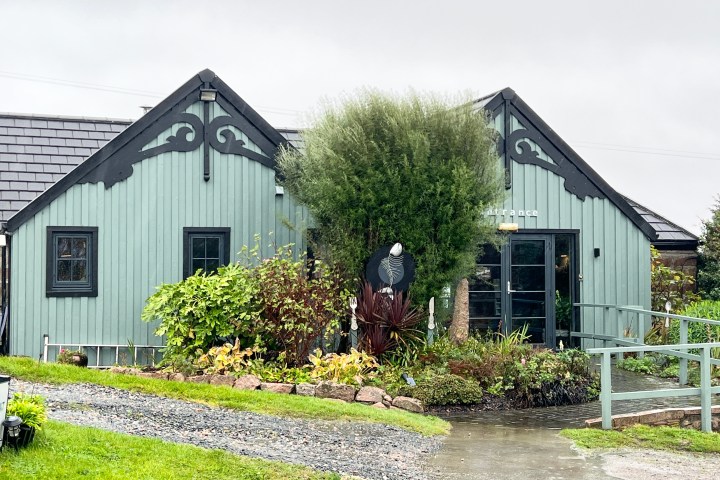A teal, wooden building with decorative trim and a landscaped entrance on a cloudy day.