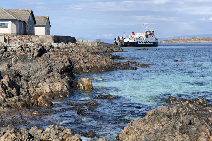 Rocky coastline with a docked ferry and coastal buildings under a blue sky.