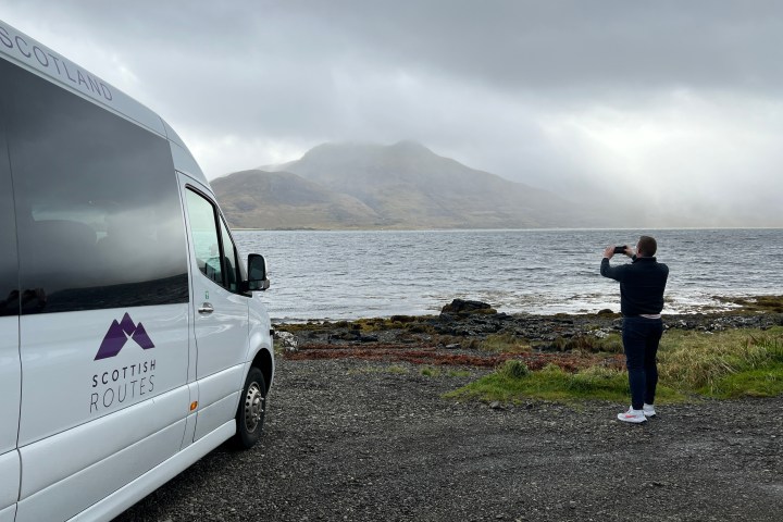 Person photographing a mountain and lake with a Scottish Routes van nearby.