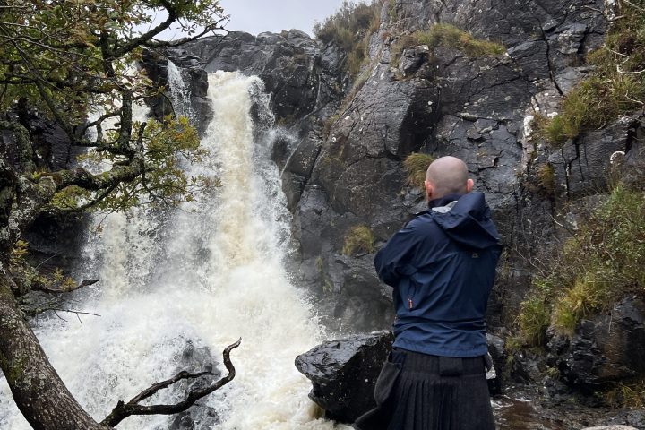 Person in kilt standing near a waterfall and rocky landscape.