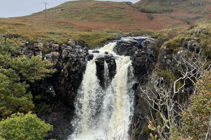 Waterfall cascading down rocks with hills and overcast sky in the background.