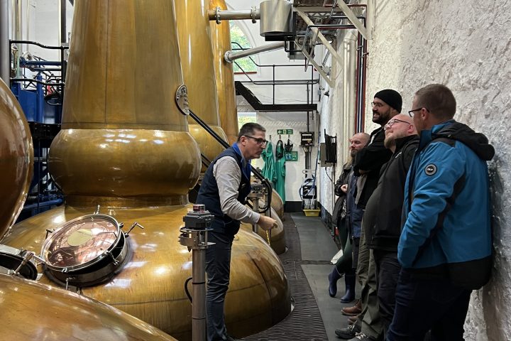 Person giving a tour inside a distillery with large copper stills and a group of people listening.
