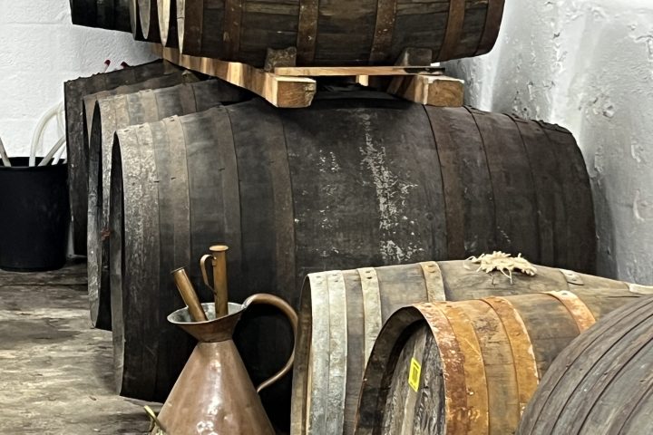Stacked wooden barrels and a copper pitcher in a white-walled cellar.