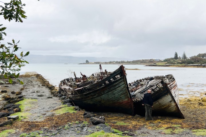 Two old, weathered boats on a rocky shore with a person standing nearby under a cloudy sky.