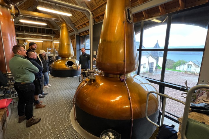 People in a distillery room with copper stills, observing and listening to a guide.