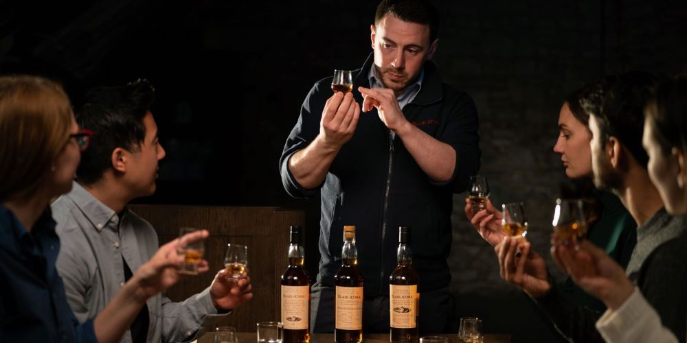 Man leading whisky tasting session; people seated with glasses and bottles on table.