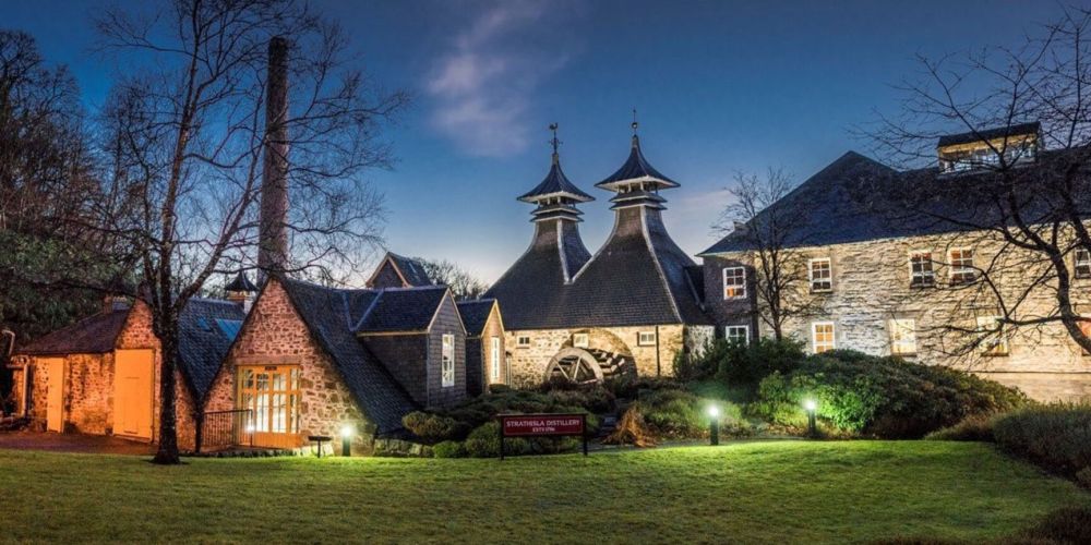 Illuminated distillery buildings with distinctive chimneys at dusk, surrounded by trees and lawn.